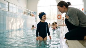 Young child wearing a neoprene swimming headband standing in the shallow end of an indoor pool, smiling at a parent kneeling at the poolside